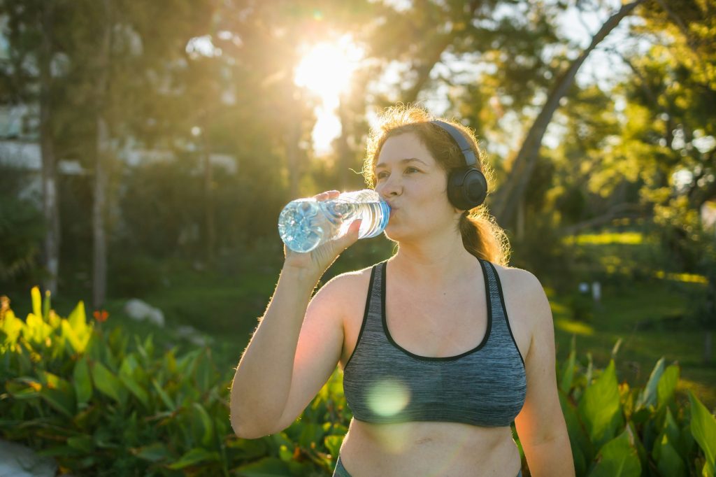 Overweight woman drinking water after jogging in the park. Portrait of young plus-size thirsty woman